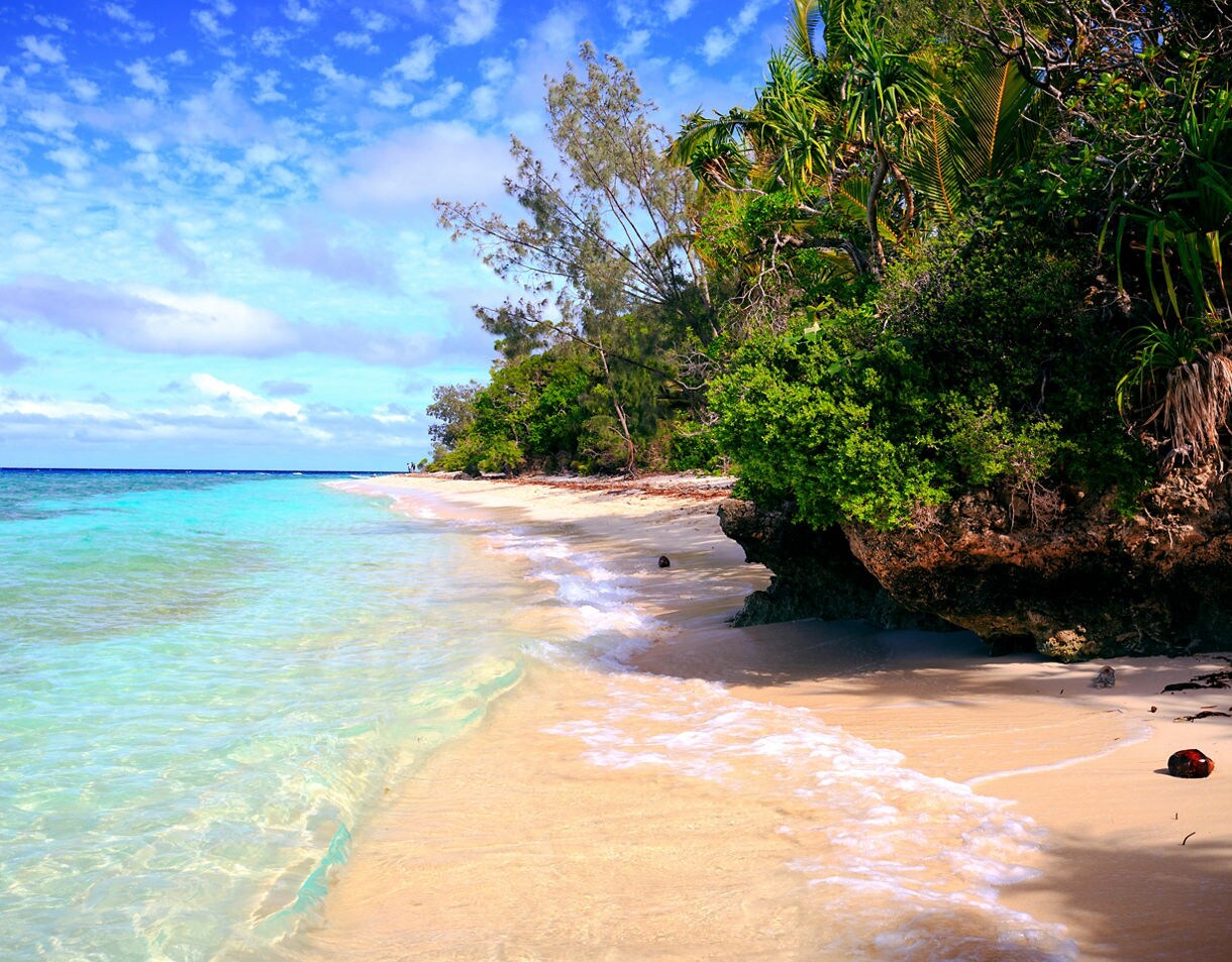 Clear waves washing over a pale sandy beach beside rocky, greenery-covered cliffs under a bright sky.
