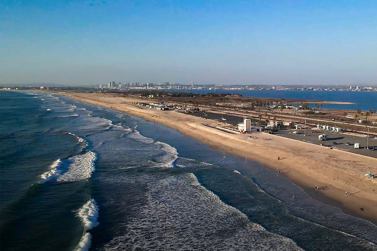 Aerial view of Silver Strand Beach in California, showcasing a long stretch of golden sand between the Pacific Ocean and San Diego Bay. Gentle waves roll onto the shore as a few beachgoers stroll along the water.
