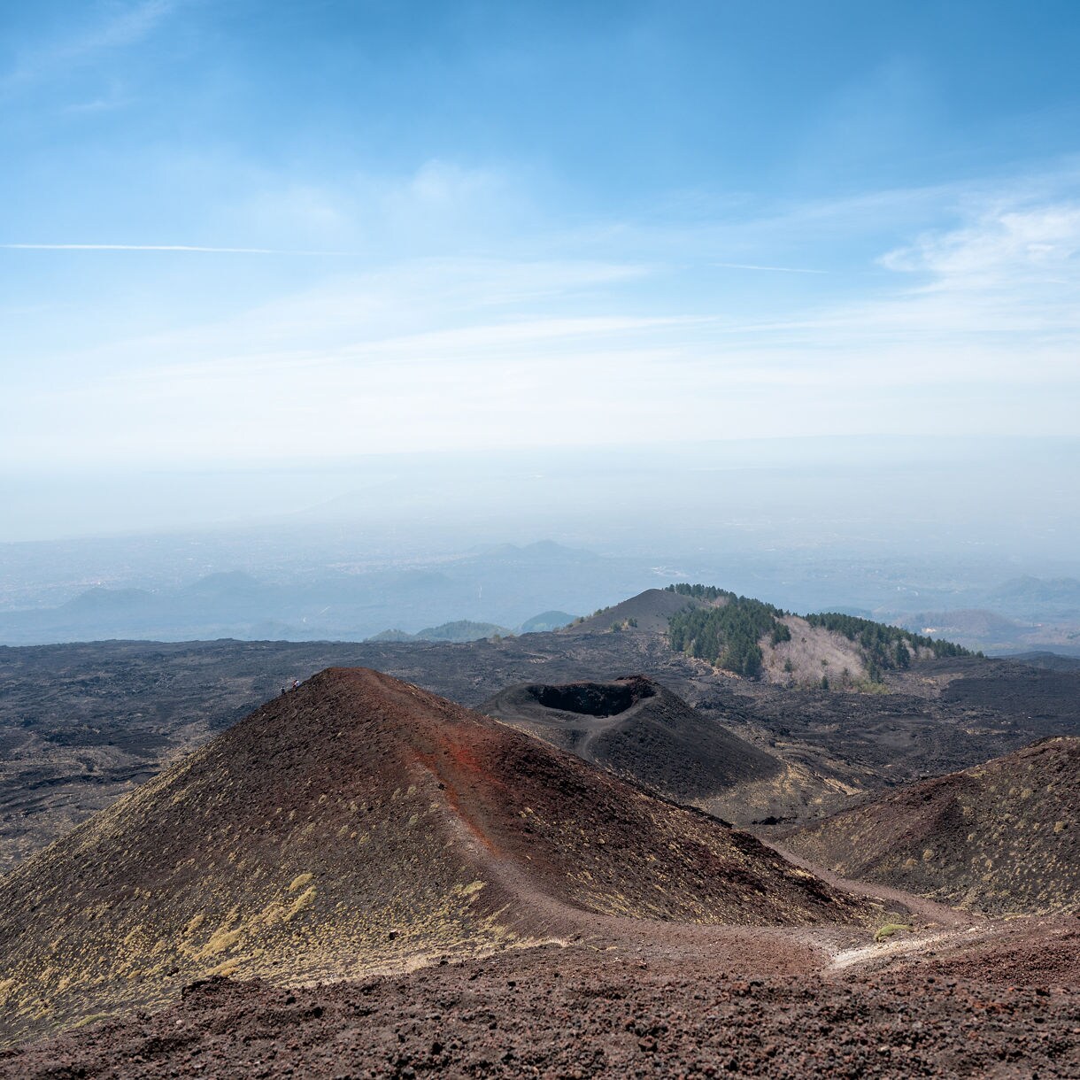 Expansive view of the Silvestri Craters on Mount Etna, showing rust-colored volcanic cones set against dark lava fields with distant hills and a hazy coastline beneath a pale blue sky.