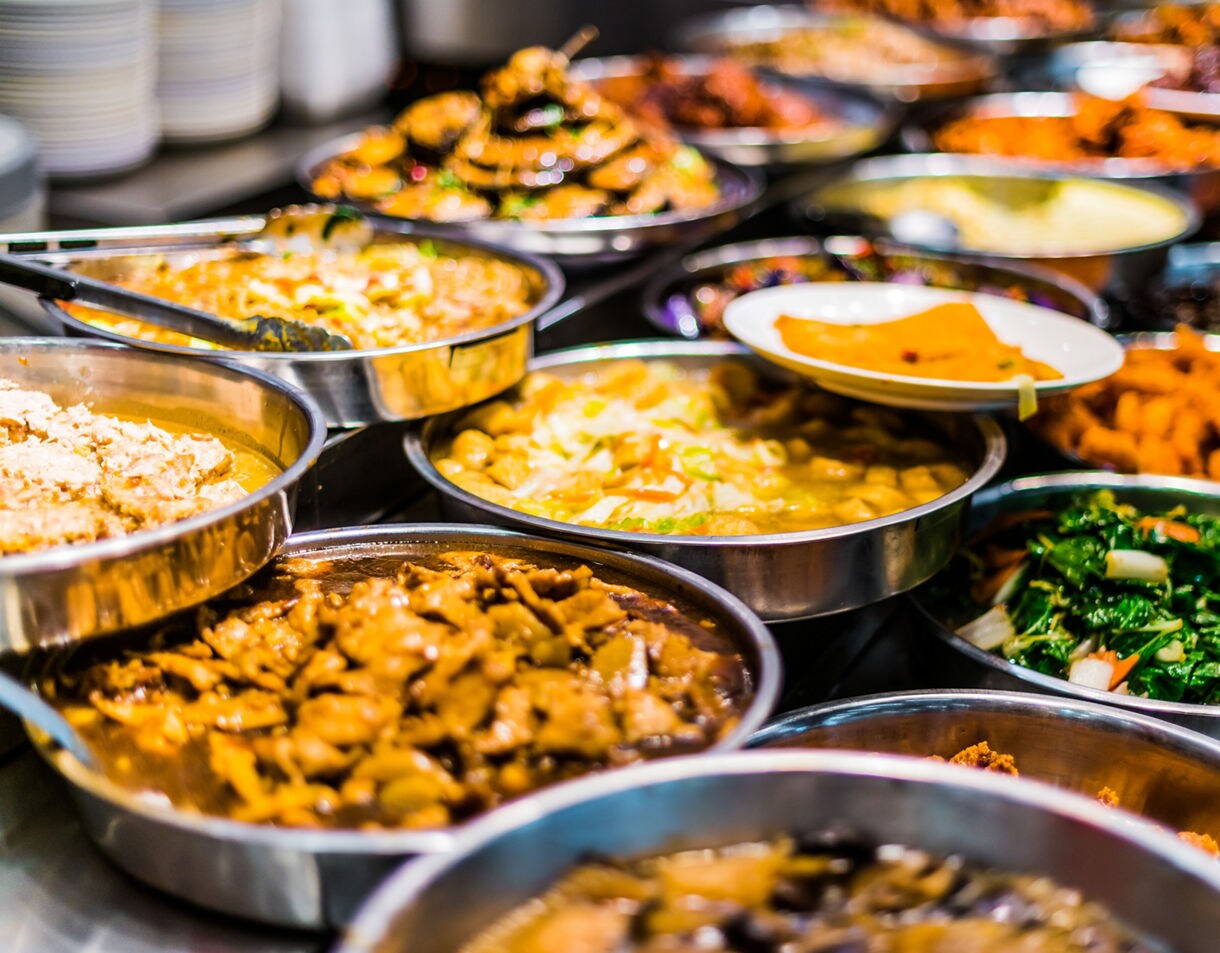 Assorted trays of colorful Singaporean street food including curries, vegetables, and stir-fried dishes displayed at a hawker stall.