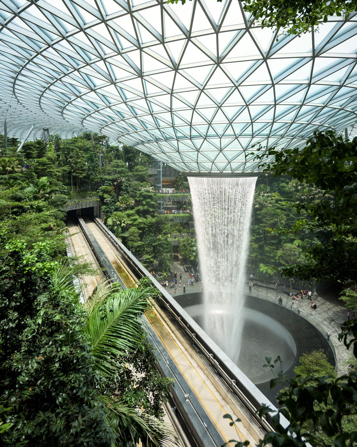 view of the Jewel Changi Airport waterfall cascading from a glass dome into a lush indoor forest with a train track nearby.