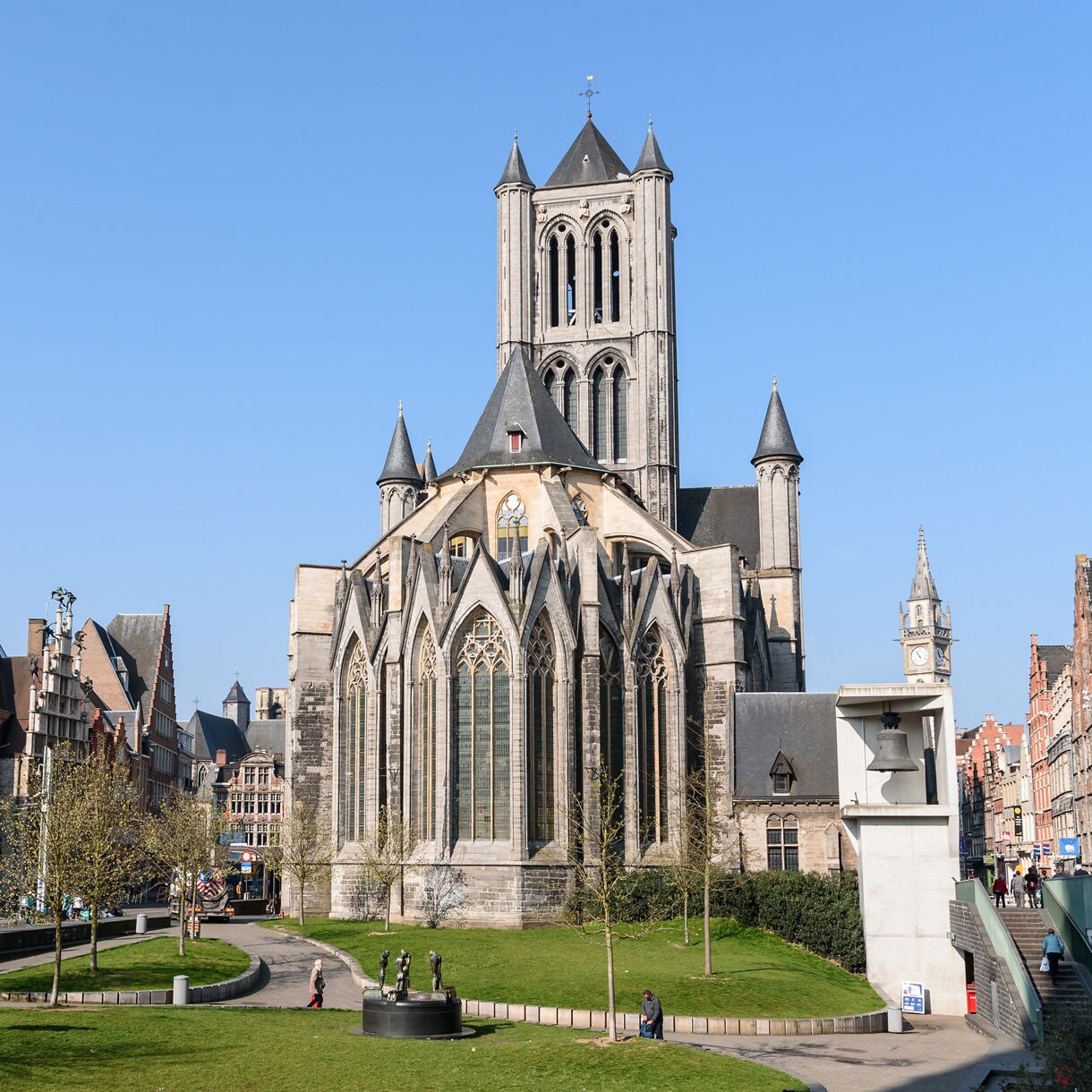 Exterior of St. Nicholas’ Church in Ghent, Belgium, featuring tall Gothic towers, pointed arches and surrounding green space with people walking nearby.