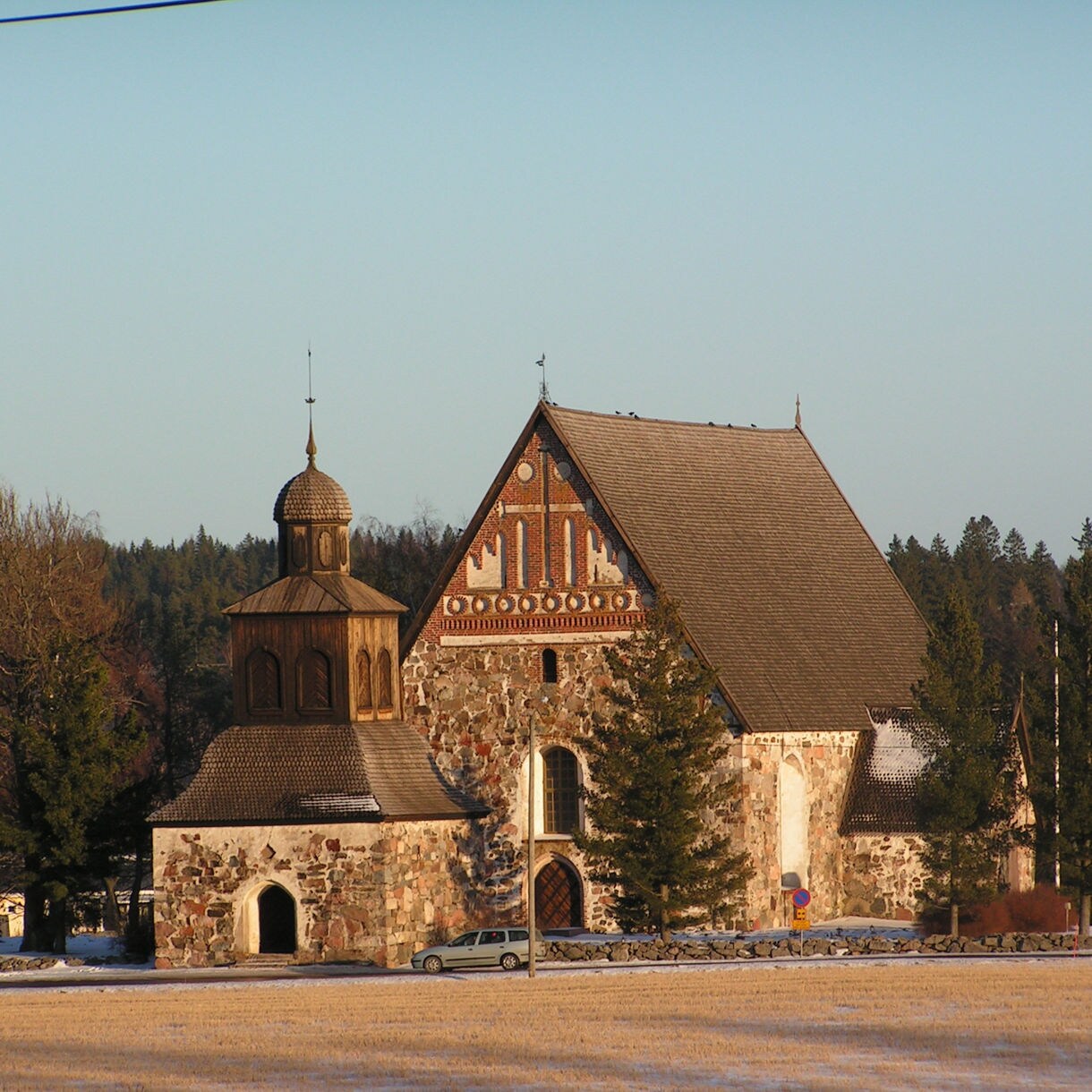 Medieval stone church in Sipoo, Finland, featuring a steep gabled roof, patterned brickwork and a wooden bell tower surrounded by trees and open fields.