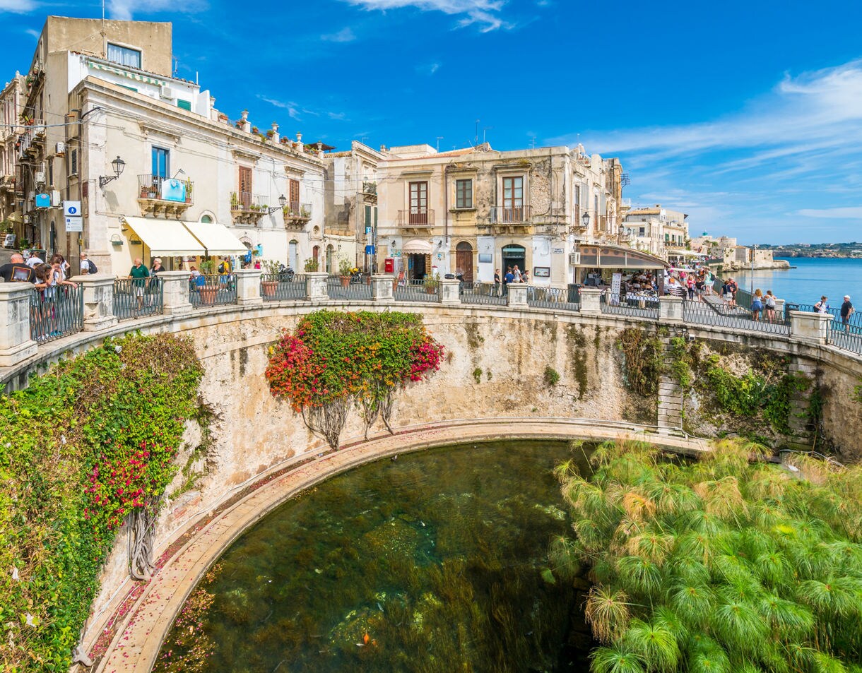 View of the Fountain of Arethusa in Siracusa, featuring a circular stone pool with lush papyrus plants and flowering vines, surrounded by historic buildings, balconies and pedestrians along the waterfront under a bright blue sky.