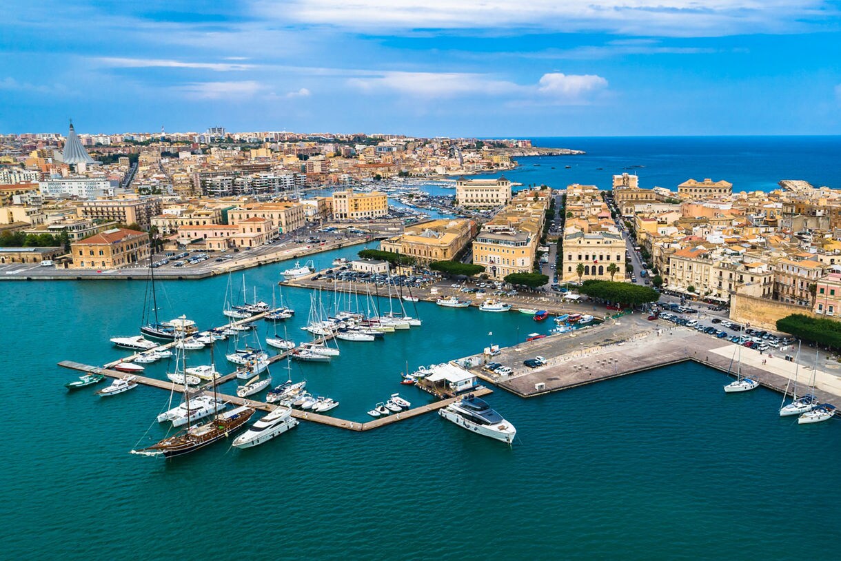 Aerial view of Siracusa, Sicily, showing a marina filled with sailboats and yachts, historic tan stone buildings packed along narrow streets and the bright blue sea stretching into the horizon under a partly cloudy sky.