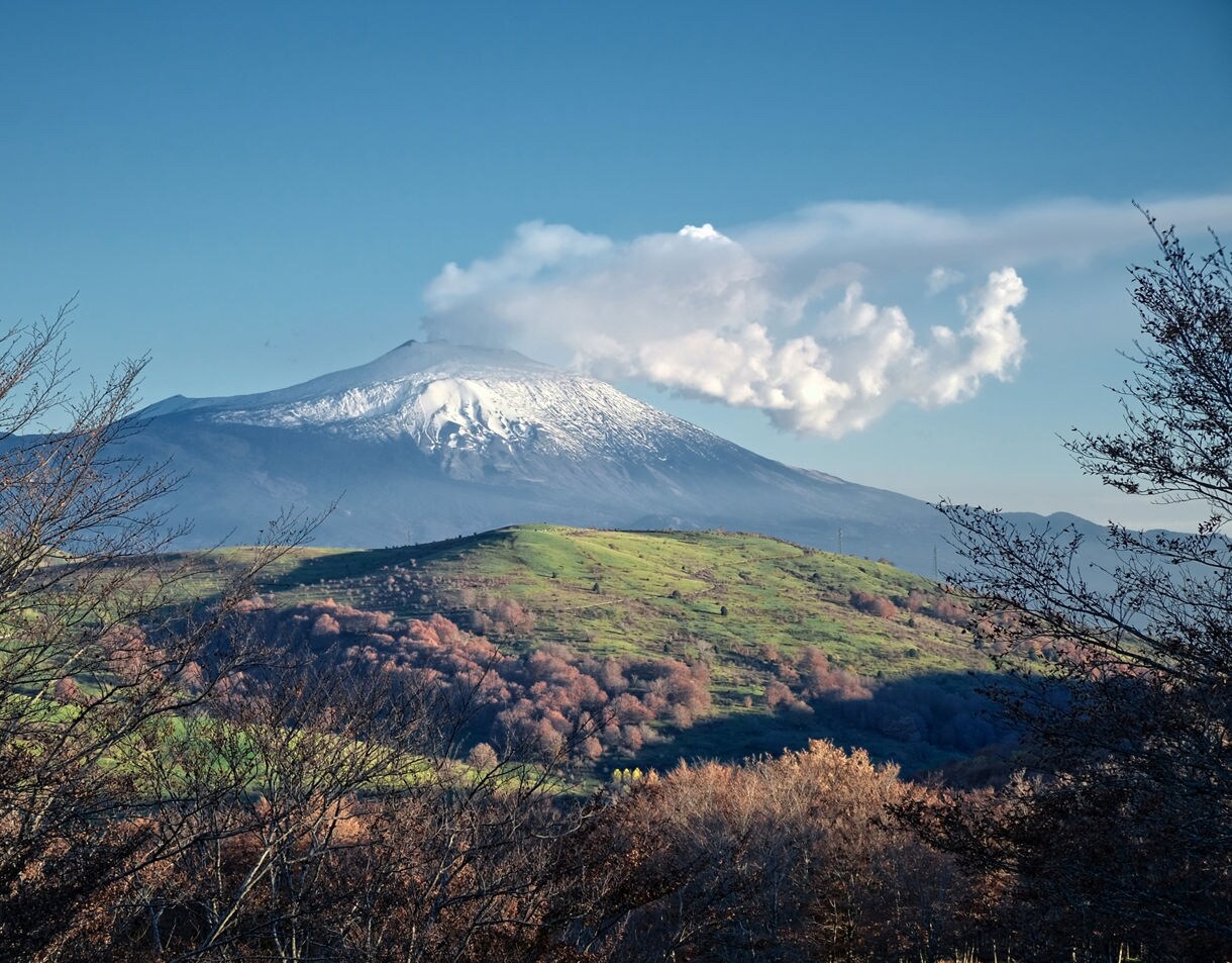 View of Mount Etna with a snow-covered summit emitting a plume of white steam, framed by rolling green and brown hills and bare winter trees under a clear blue sky.