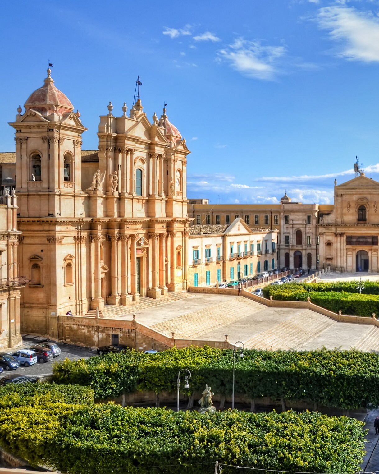 Elevated view of Noto, Sicily, showing ornate Baroque churches and palaces built from warm sandstone, a large staircase leading to a central piazza, tree-lined gardens and narrow streets under a bright blue sky.