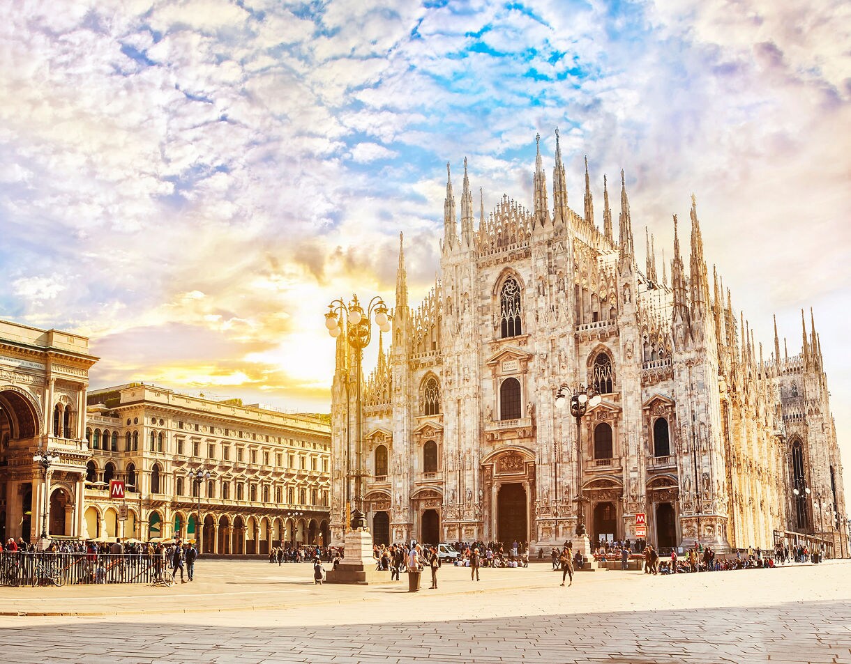 Wide view of the Piazza del Duomo in Milan featuring the ornate white marble cathedral with its many spires, the grand Galleria Vittorio Emanuele II entrance on the left and crowds of people gathered across the sunlit square under a colorful sky.