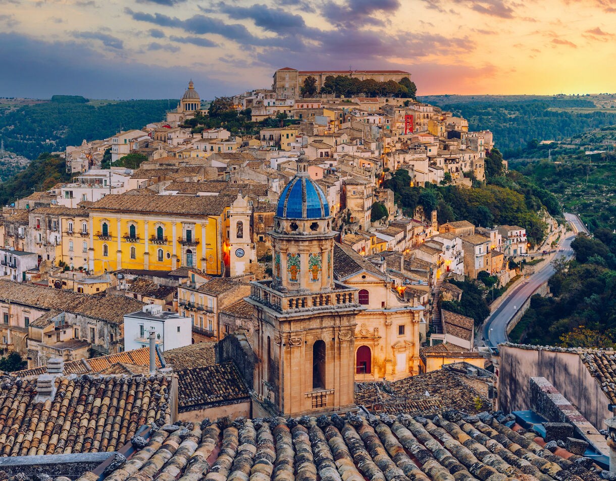 Panoramic view of Ragusa Ibla in Sicily at sunset featuring a blue-domed church tower, clusters of stone buildings with tile roofs, terraced streets and surrounding green valleys under a warm, colorful sky.