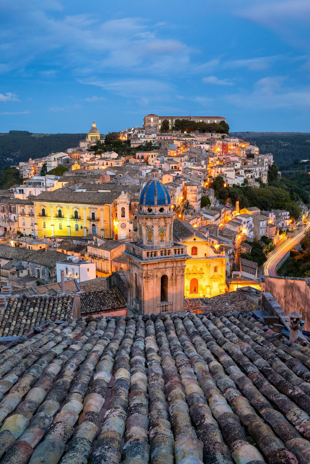Vertical view of Ragusa Ibla showing terracotta rooftops in the foreground, a blue-domed church tower and clusters of illuminated stone buildings cascading down a hill, with surrounding green valleys under a twilight sky.