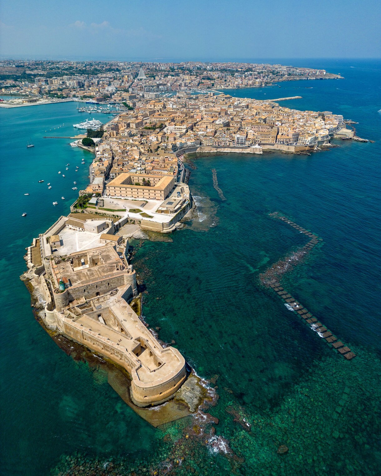 Aerial vertical view of Ortigia Island in Siracusa, featuring the coastal fortress of Castello Maniace in the foreground, dense historic buildings in tan stone, a marina with boats and vivid turquoise water surrounding the peninsula under a clear blue sky.