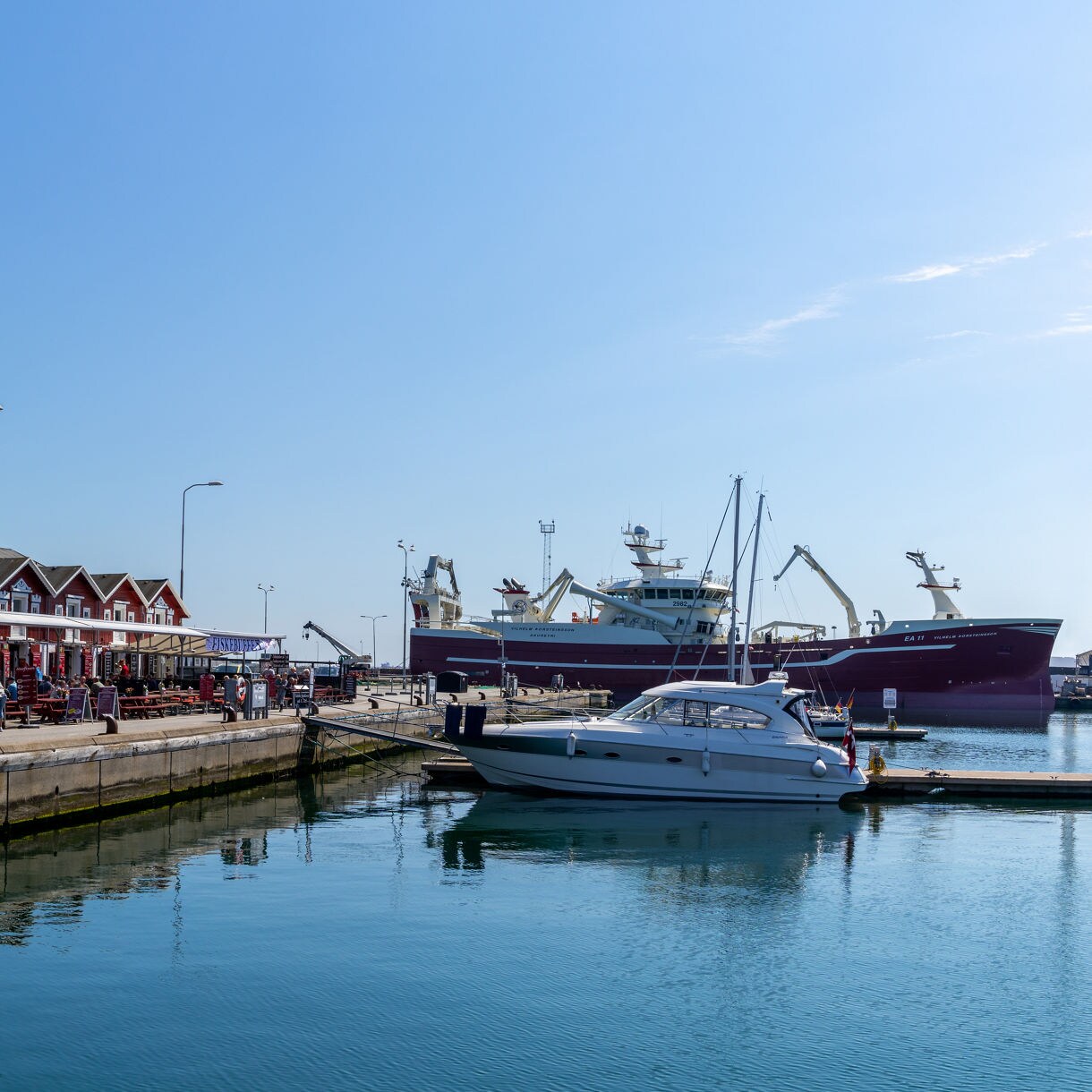 Skagen Harbor with red wooden buildings, outdoor restaurants, fishing boats and a large ship docked nearby.
