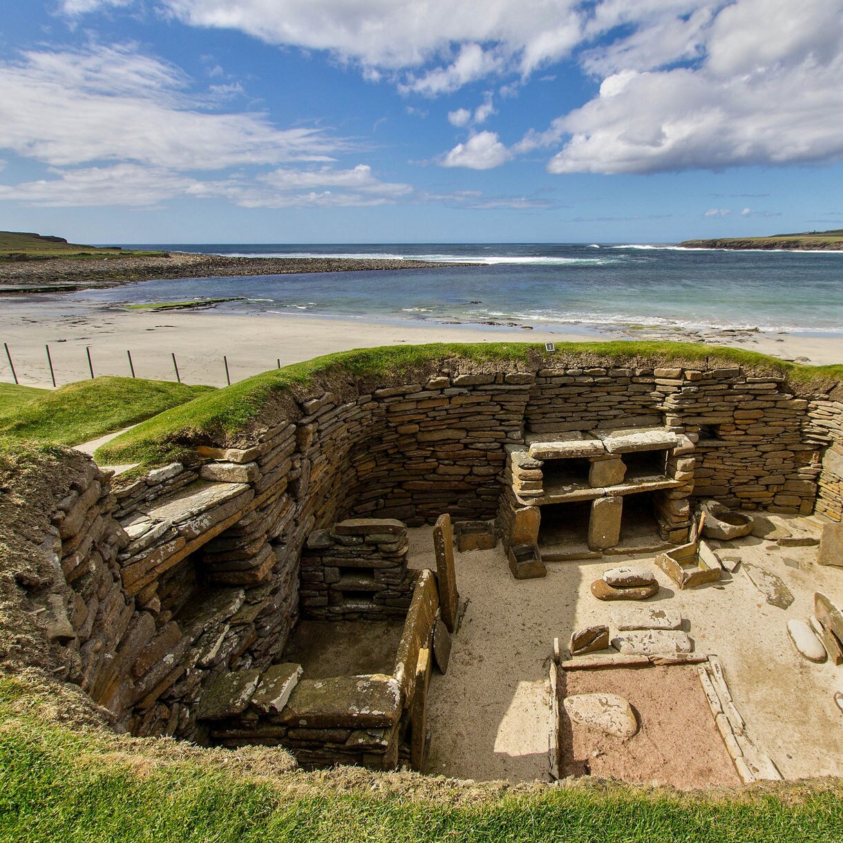 Neolithic stone houses of Skara Brae in Orkney, with intact stone walls and hearths overlooking a sandy beach and the Atlantic Ocean.