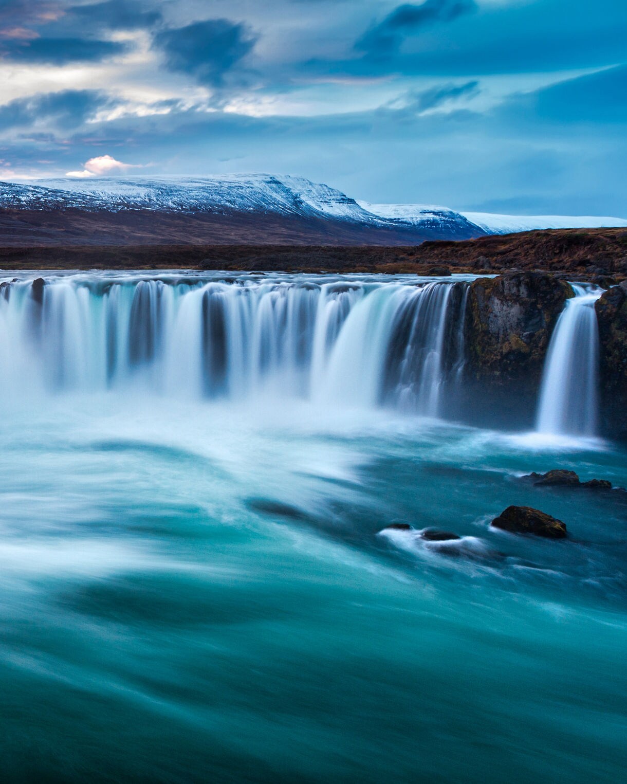Goðafoss waterfall in Iceland, with wide cascades of turquoise water plunging into a swirling river, framed by rocky cliffs and distant snow-capped mountains under a dramatic sky.