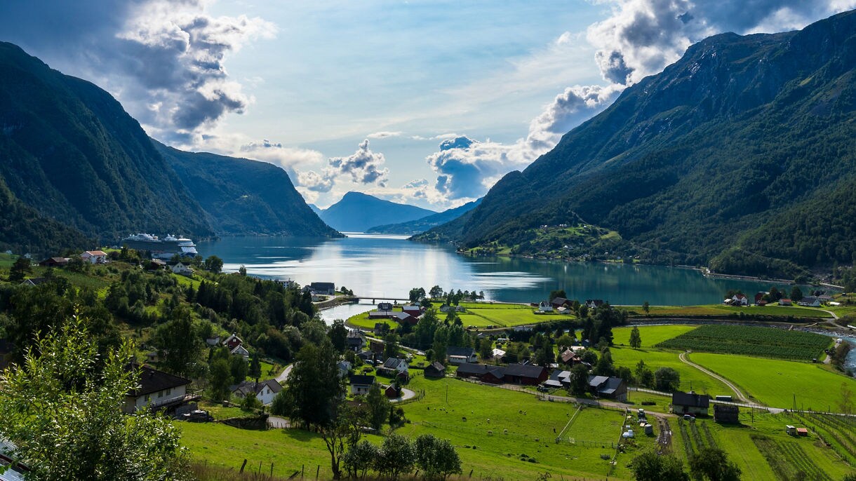 A panoramic view of Skjolden, Norway, with green farmland and scattered houses leading to a reflective fjord surrounded by steep, forested mountains under a dramatic sky.