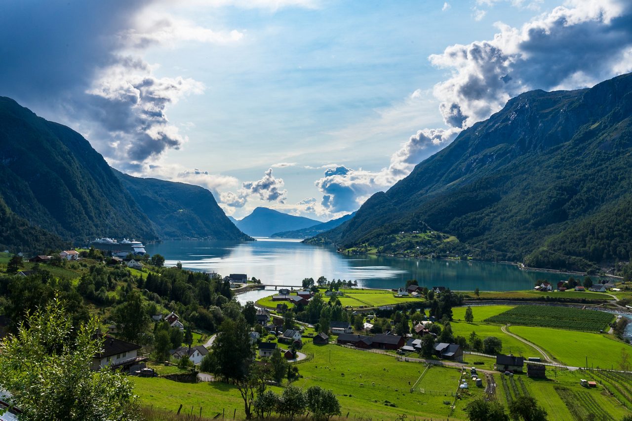 A panoramic view of Skjolden, Norway, with green farmland and scattered houses leading to a reflective fjord surrounded by steep, forested mountains under a dramatic sky.