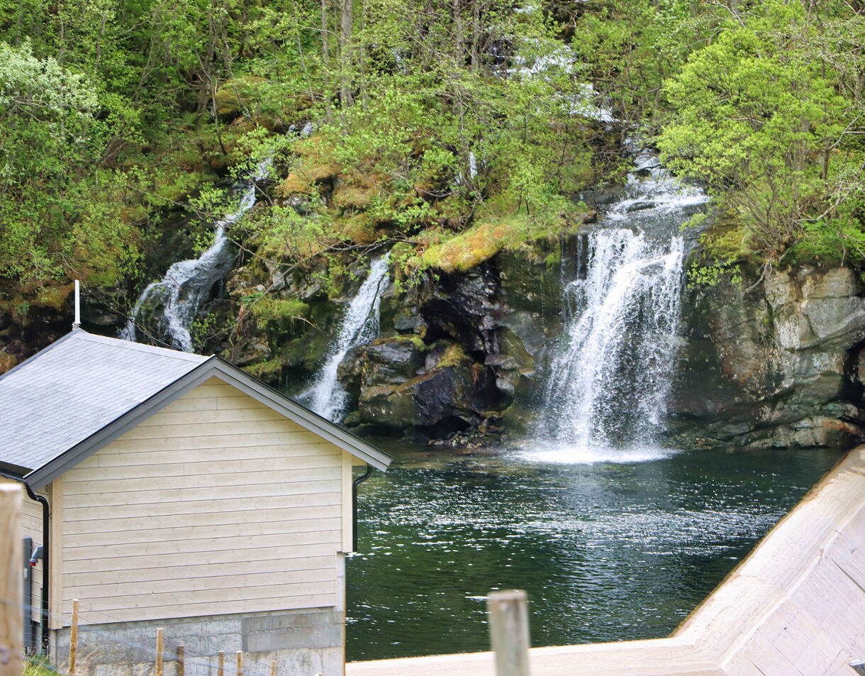 Twin waterfalls cascade down mossy rock cliffs into a calm pool in Skjolden, Norway, with a small house and dam structure in the foreground surrounded by greenery.