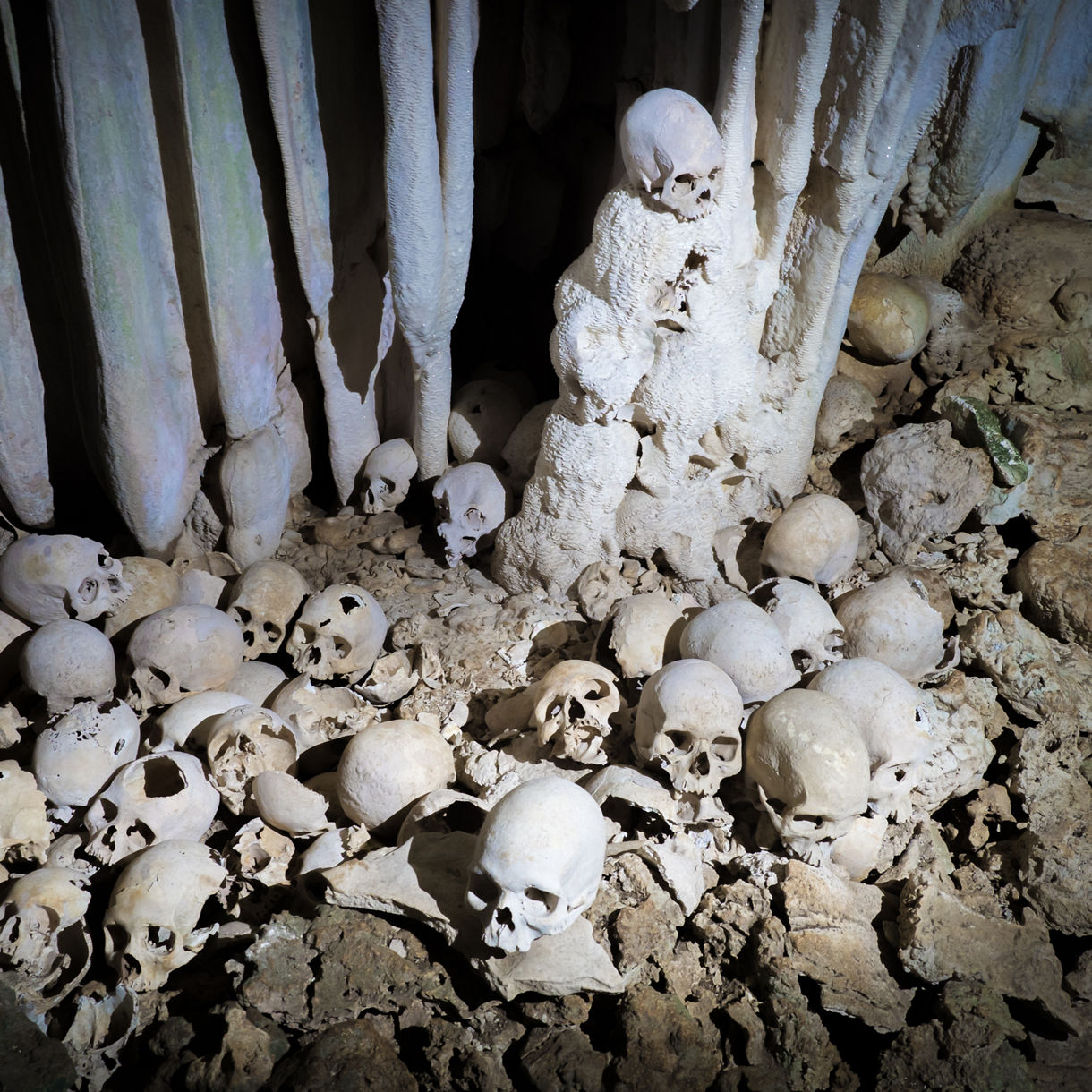 Dimly lit cave chamber lined with pale stalactites and stalagmites, surrounded by numerous human skulls and bone fragments scattered across the rocky floor.