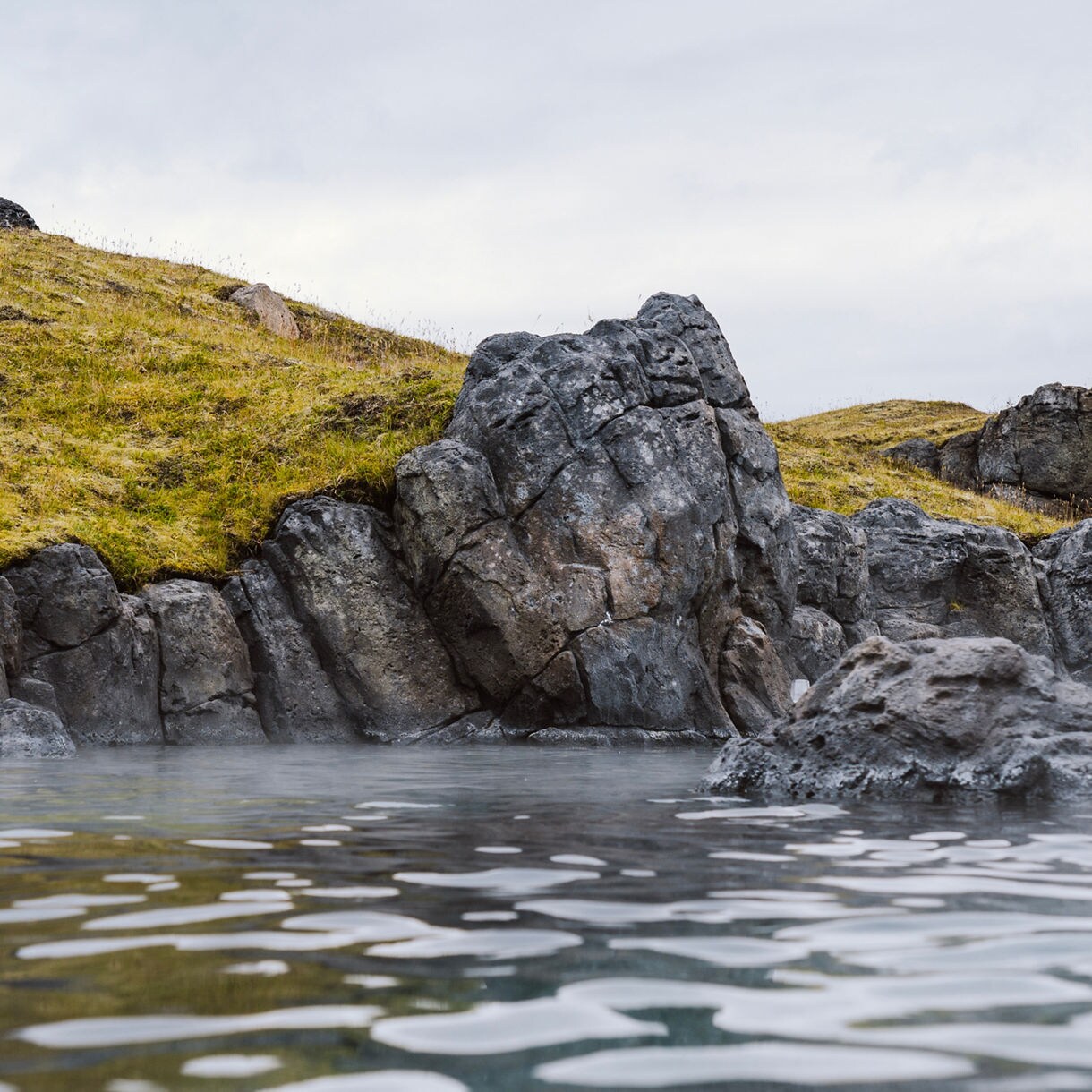 Geothermal pool at Sky Lagoon in Iceland, with steam rising from calm water surrounded by rocky formations and grassy hillsides under a cloudy sky.