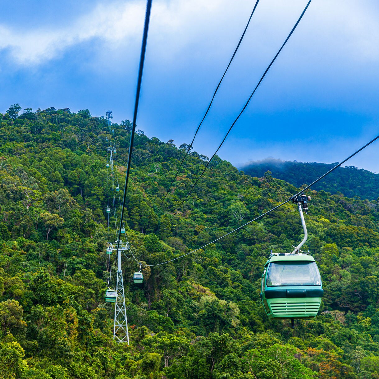 Green cable cars traveling over dense tropical rainforest beneath a cloudy blue sky in Kuranda, Queensland.