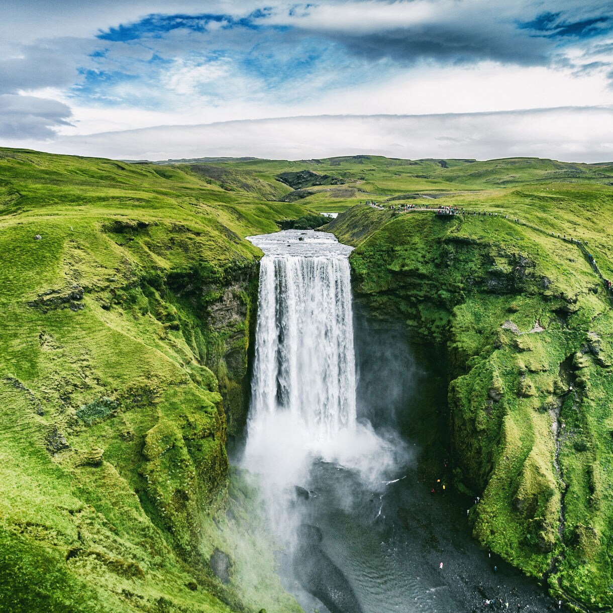 Skógafoss waterfall in Iceland, a tall curtain of water dropping into a misty pool, framed by steep green moss-covered cliffs under a partly cloudy sky.