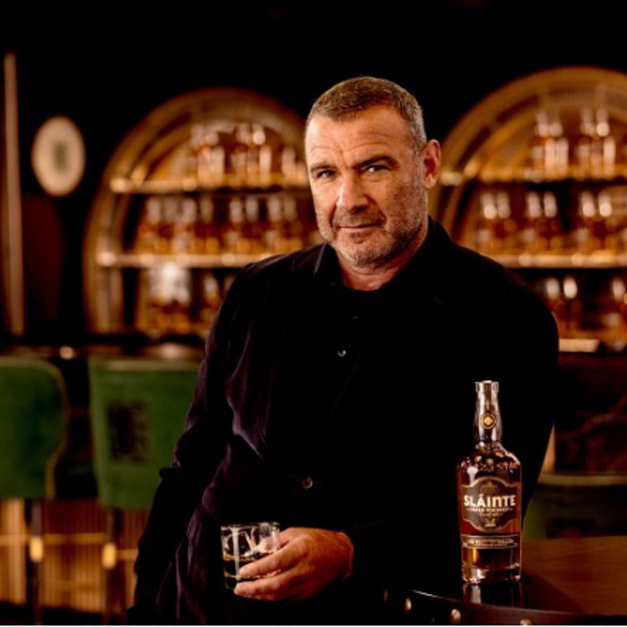 Liev Schreiber with a glass and a bottle of Slainte Irish Whisky at the bar.