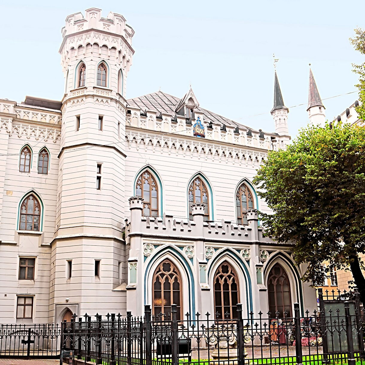 The Small Guild House in Riga, Latvia, featuring white stone walls, pointed arched windows, decorative battlements and a round tower with spires framed by trees and iron fencing.