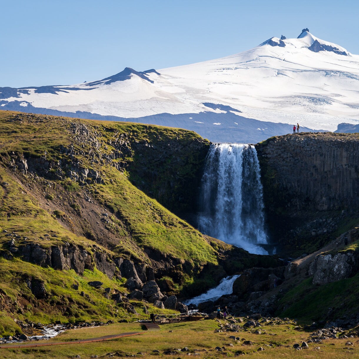 A wide waterfall flowing over a cliff of basalt columns, set in green rolling hills, with the snow-covered Snæfellsjökull volcano towering in the background under a clear blue sky.