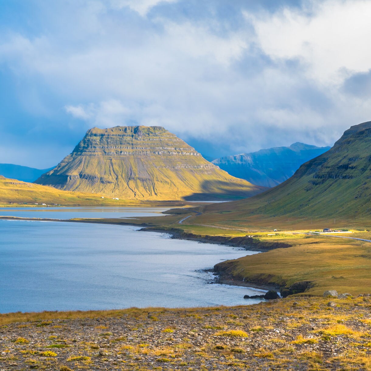 A wide valley with a calm fjord on the left and a distinct pyramid-like mountain ahead, with green and yellow slopes lit by patches of sunlight under a cloudy sky.
