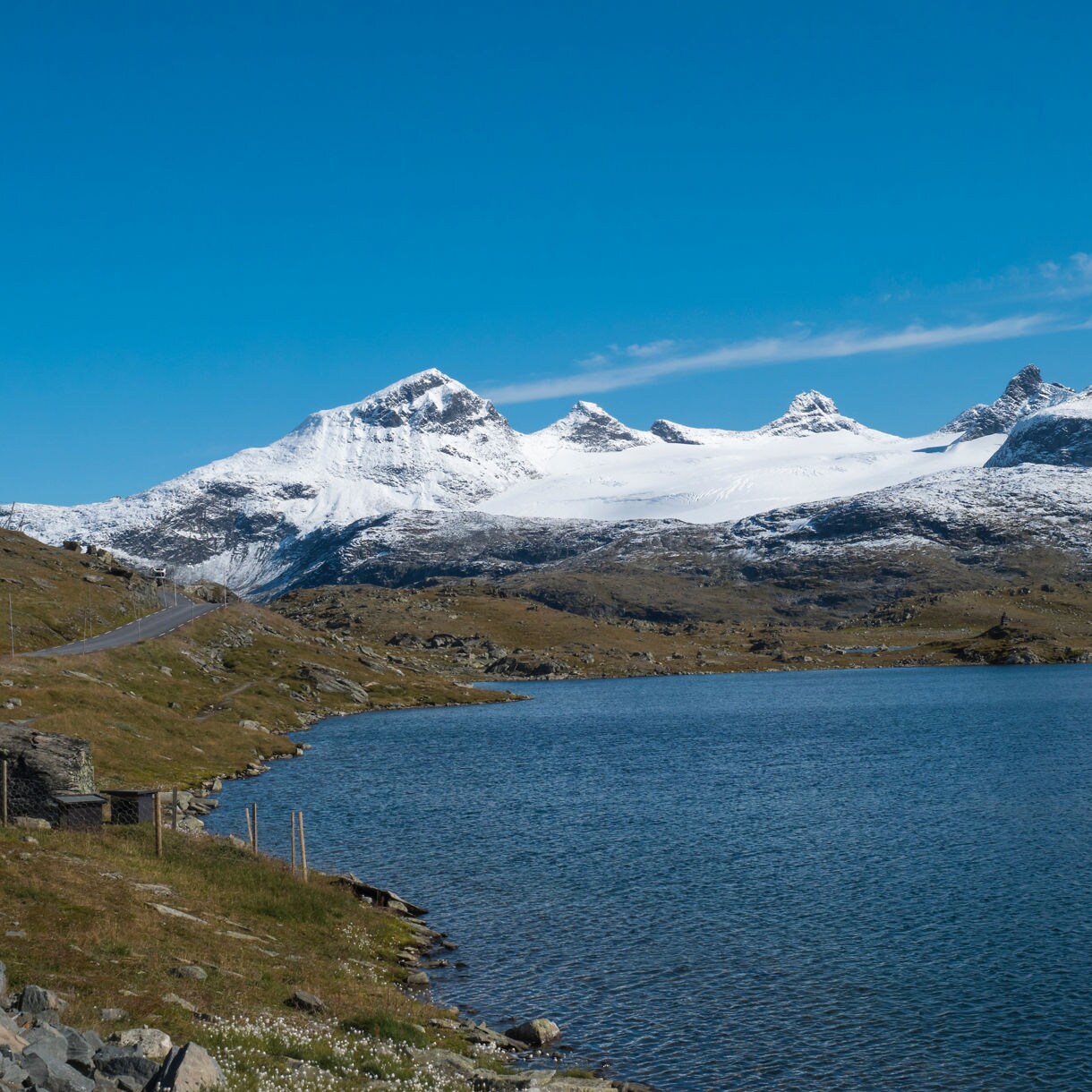 Panoramic view of Sognefjell in Norway, featuring a winding road, blue alpine lake and snow-covered mountain peaks under a bright blue sky.