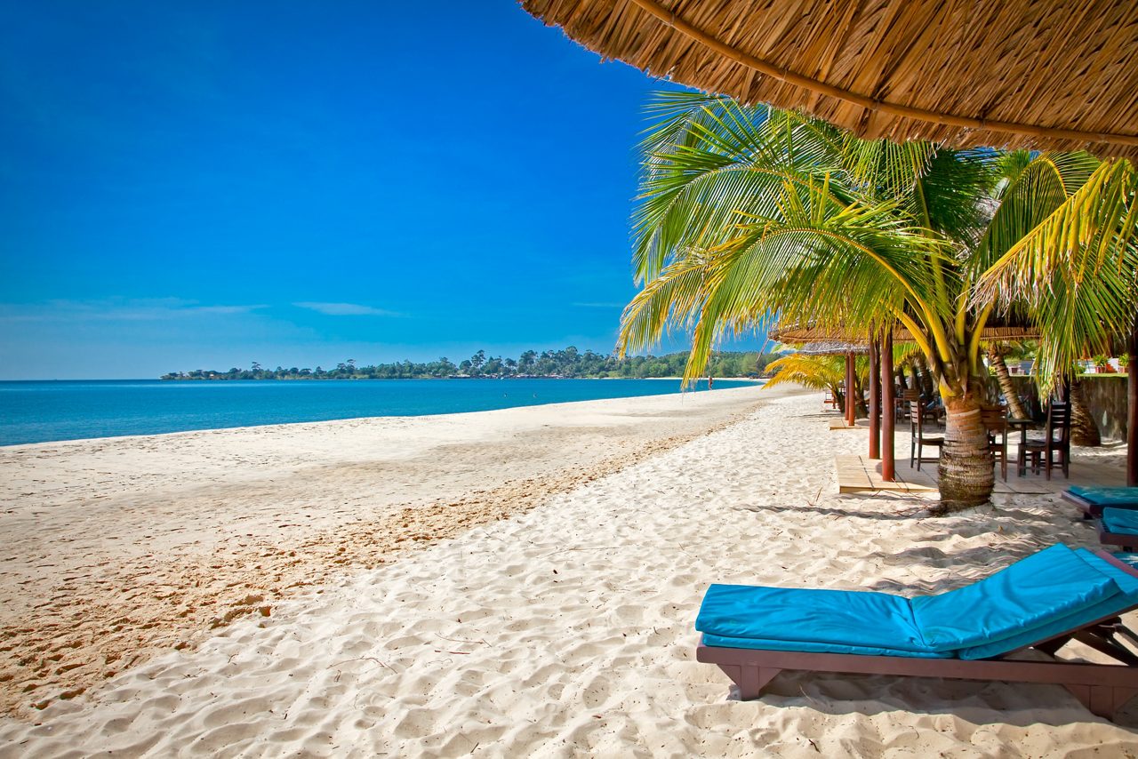 A peaceful white-sand beach with turquoise water, palm trees and shaded lounge chairs under a bright blue sky.
