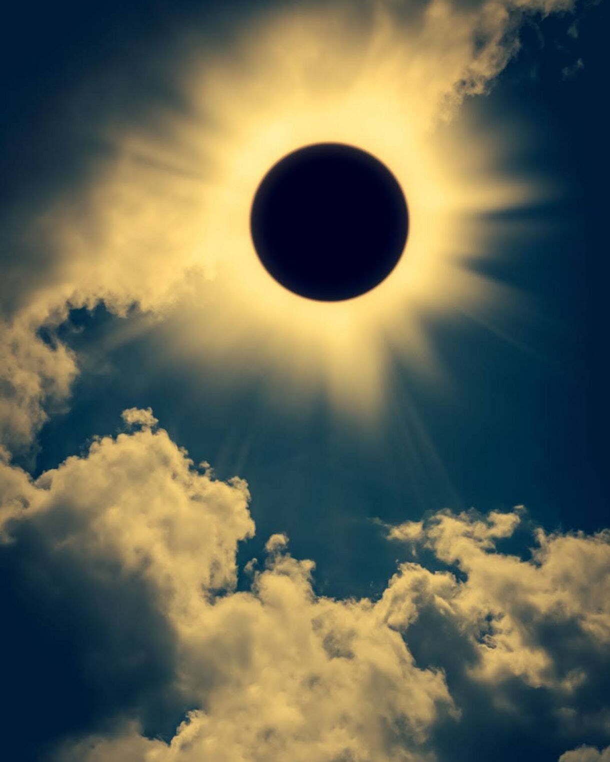 A total solar eclipse with the dark moon centered against a glowing golden corona, surrounded by dramatic clouds in a deep blue sky.