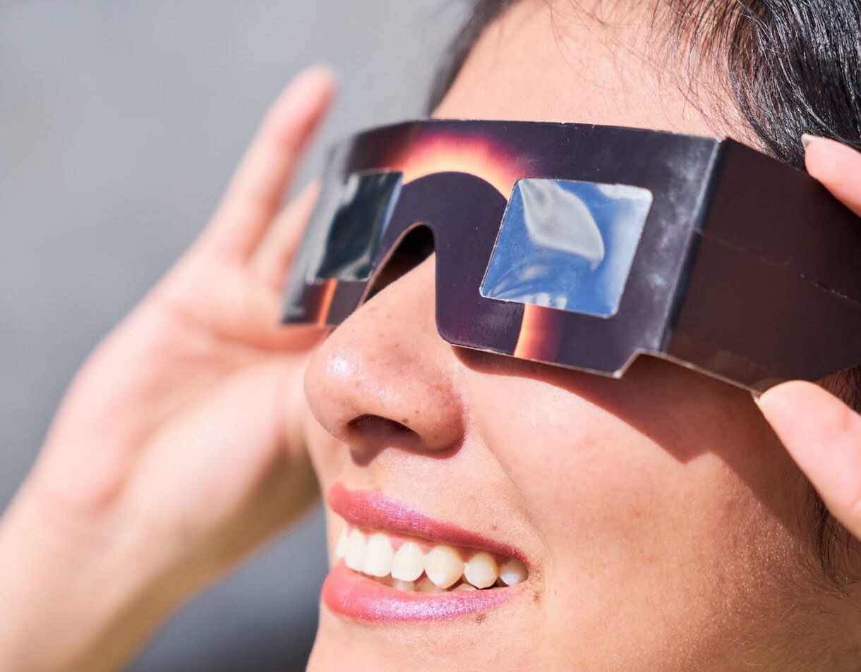 Close-up of a person smiling while wearing eclipse glasses with reflective lenses.