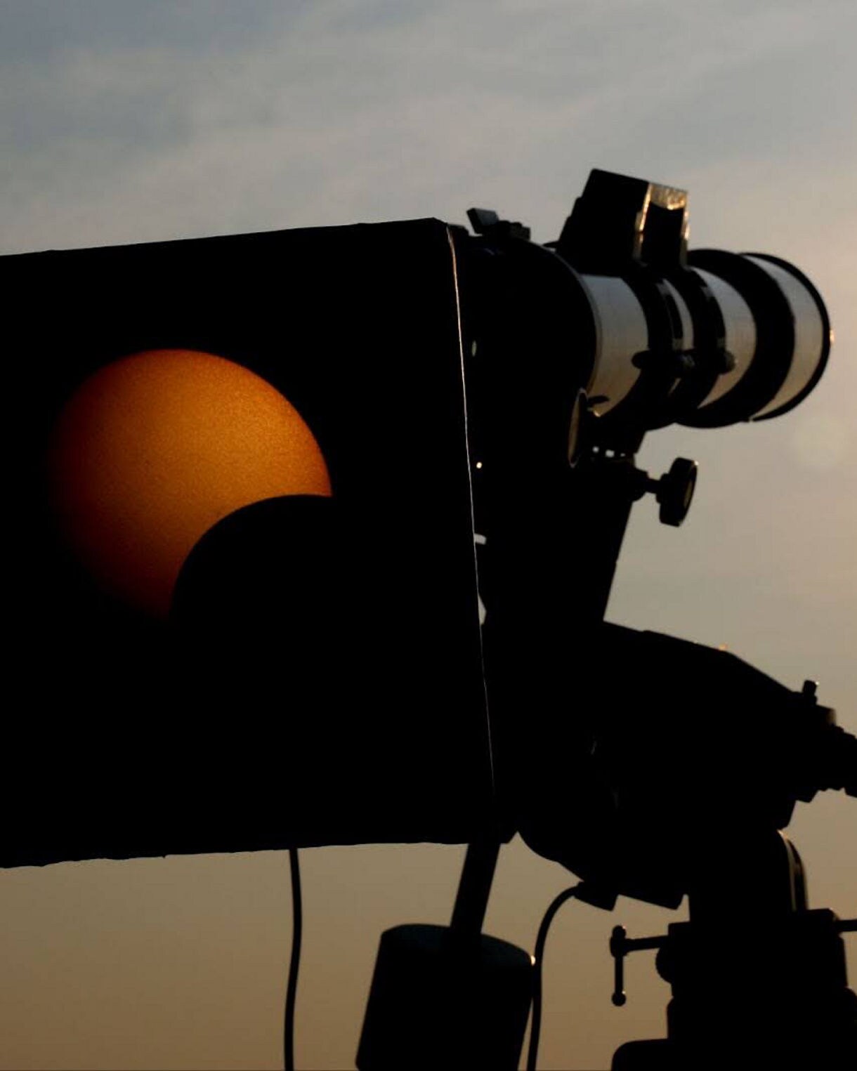 Silhouette of a telescope projecting a partial solar eclipse onto a small screen at sunset.
