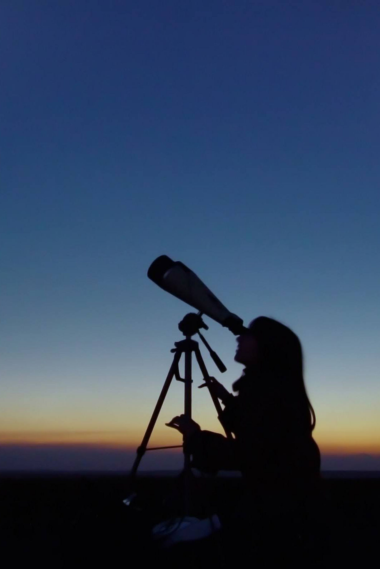 Silhouette of a person using a telescope on a tripod at dusk, watching the sky as twilight colors fade along the horizon.