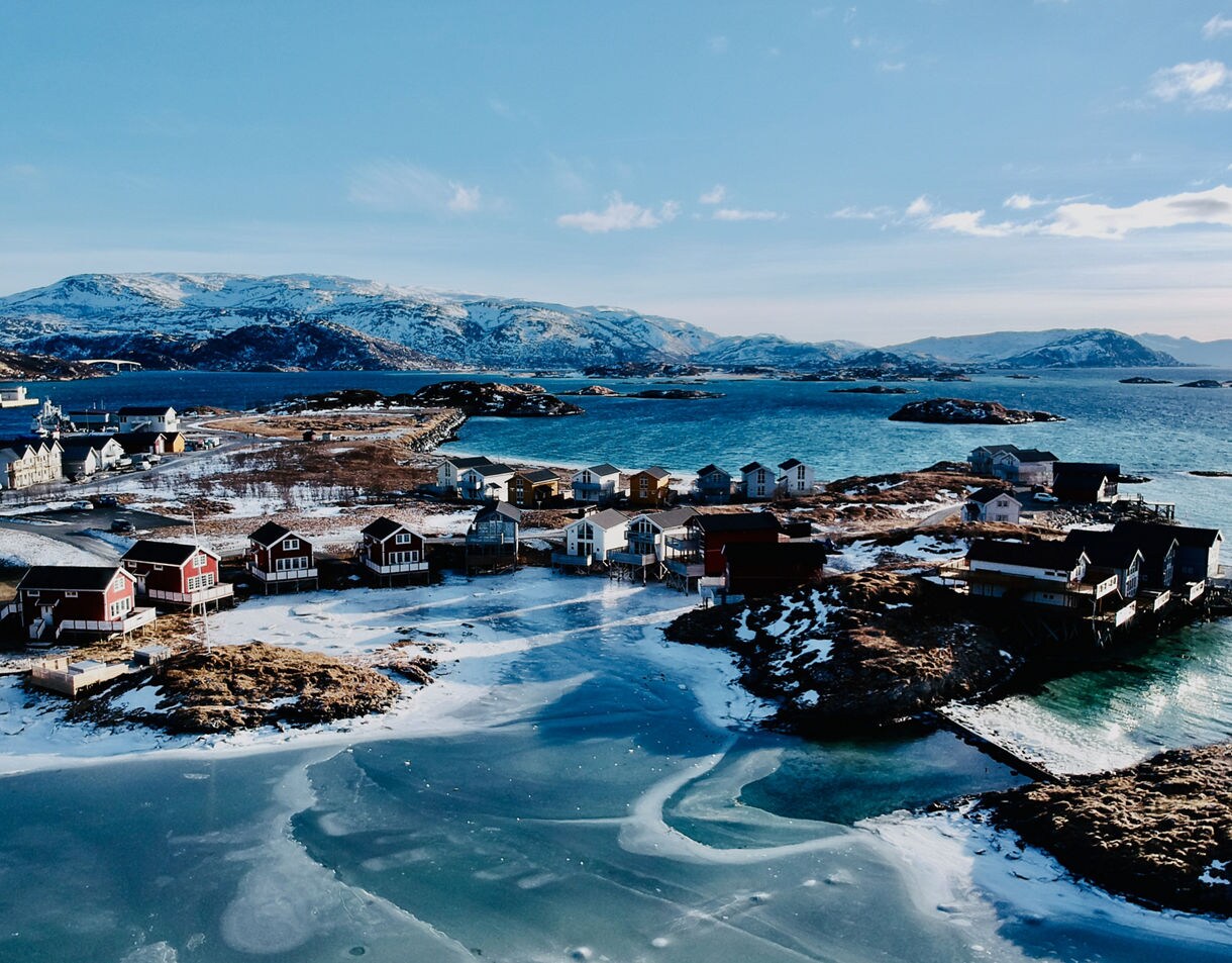 Aerial view of Sommarøy Island with red and white houses along the frozen shoreline, surrounded by blue sea and snow-covered mountains.