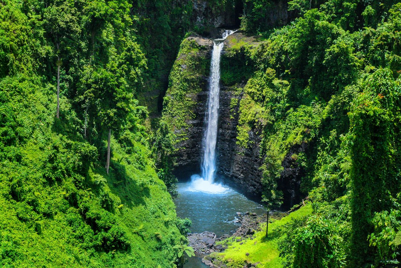 Tall waterfall plunging into a pool surrounded by steep cliffs and dense green vegetation in Samoa’s rainforest.