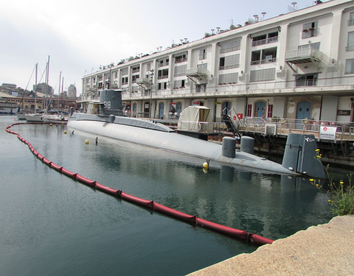 he Sottomarino Nazario Sauro submarine docked in Genoa’s Old Harbor beside white waterfront buildings and sailboats.