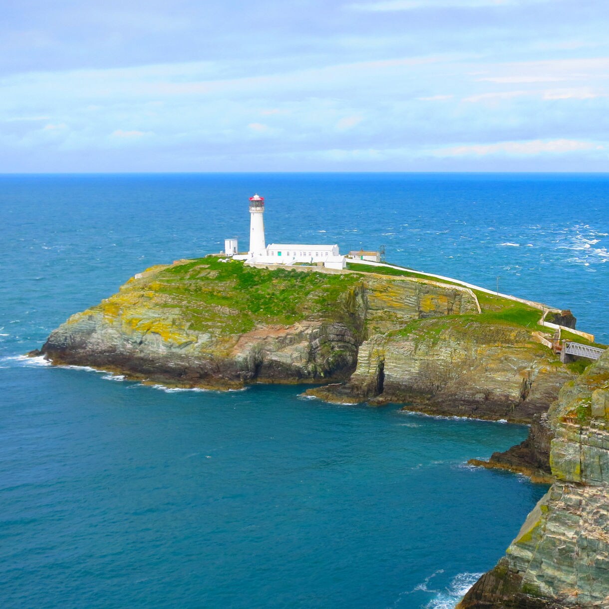White lighthouse perched on a rocky island surrounded by bright blue ocean and steep coastal cliffs.