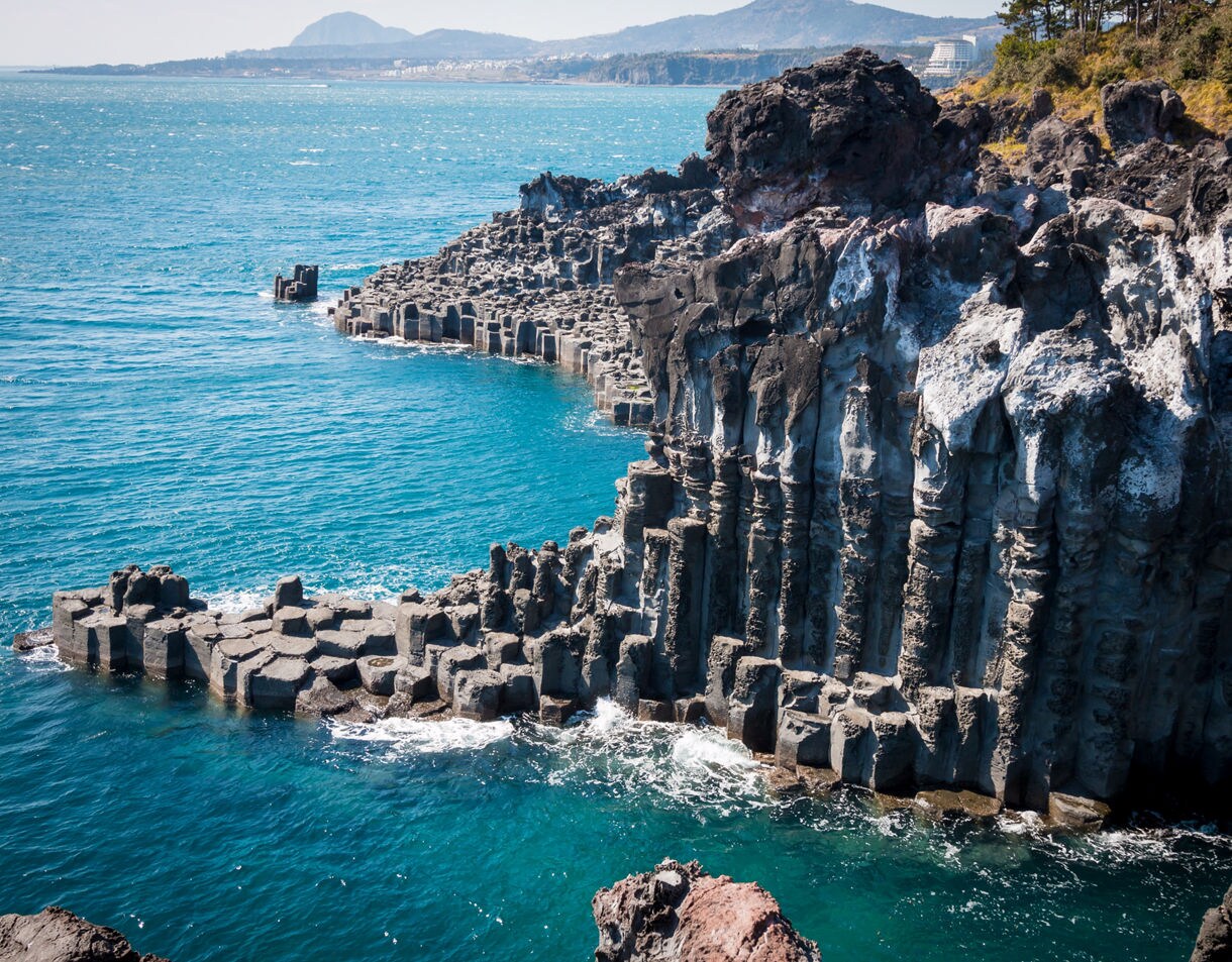 Dramatic basalt column cliffs at Jusangjeolli on Jeju Island, South Korea, rising above clear blue ocean waters with waves breaking at their base.