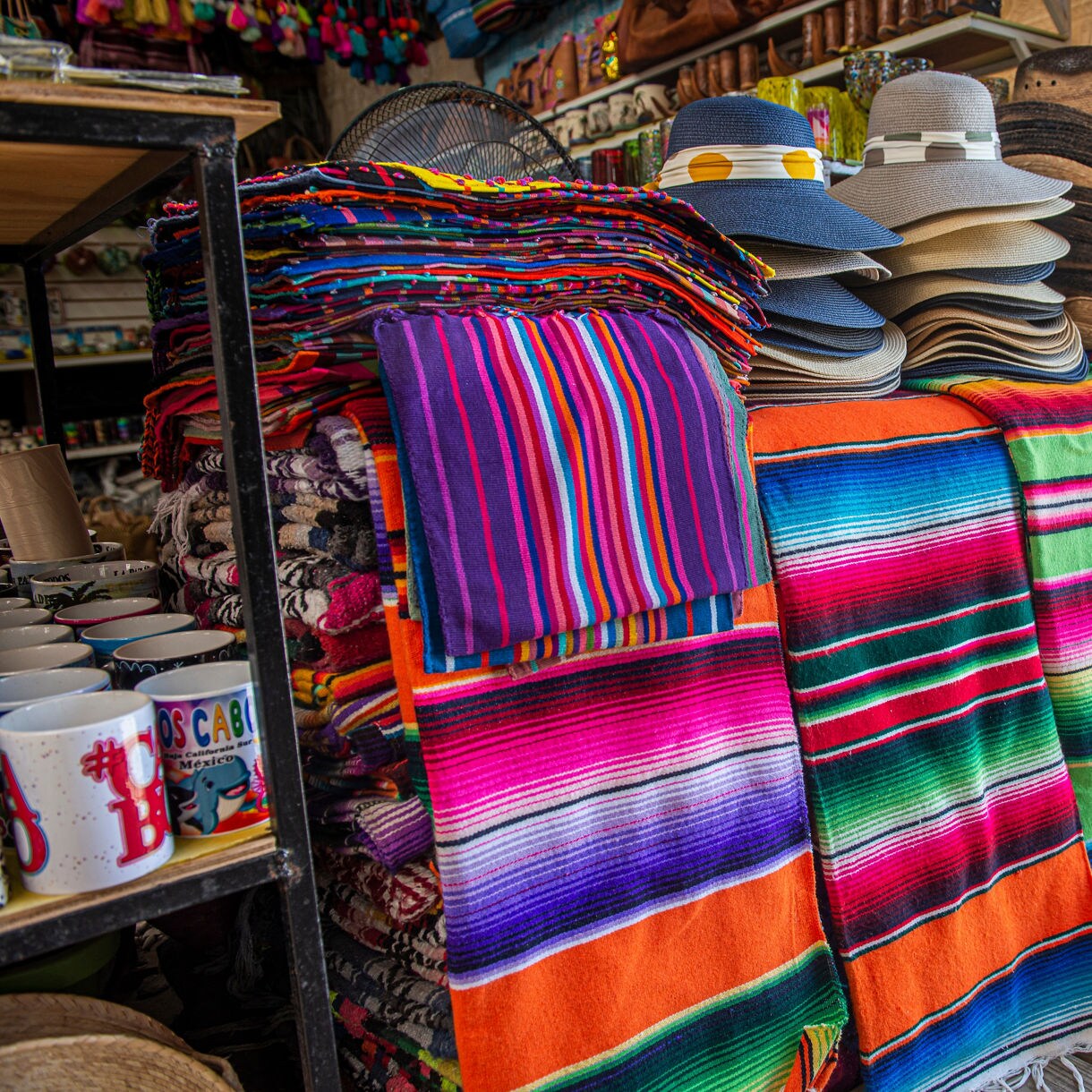 Vibrant Mexican sarapes stacked and hanging beside straw hats and mugs at a souvenir shop in La Paz.