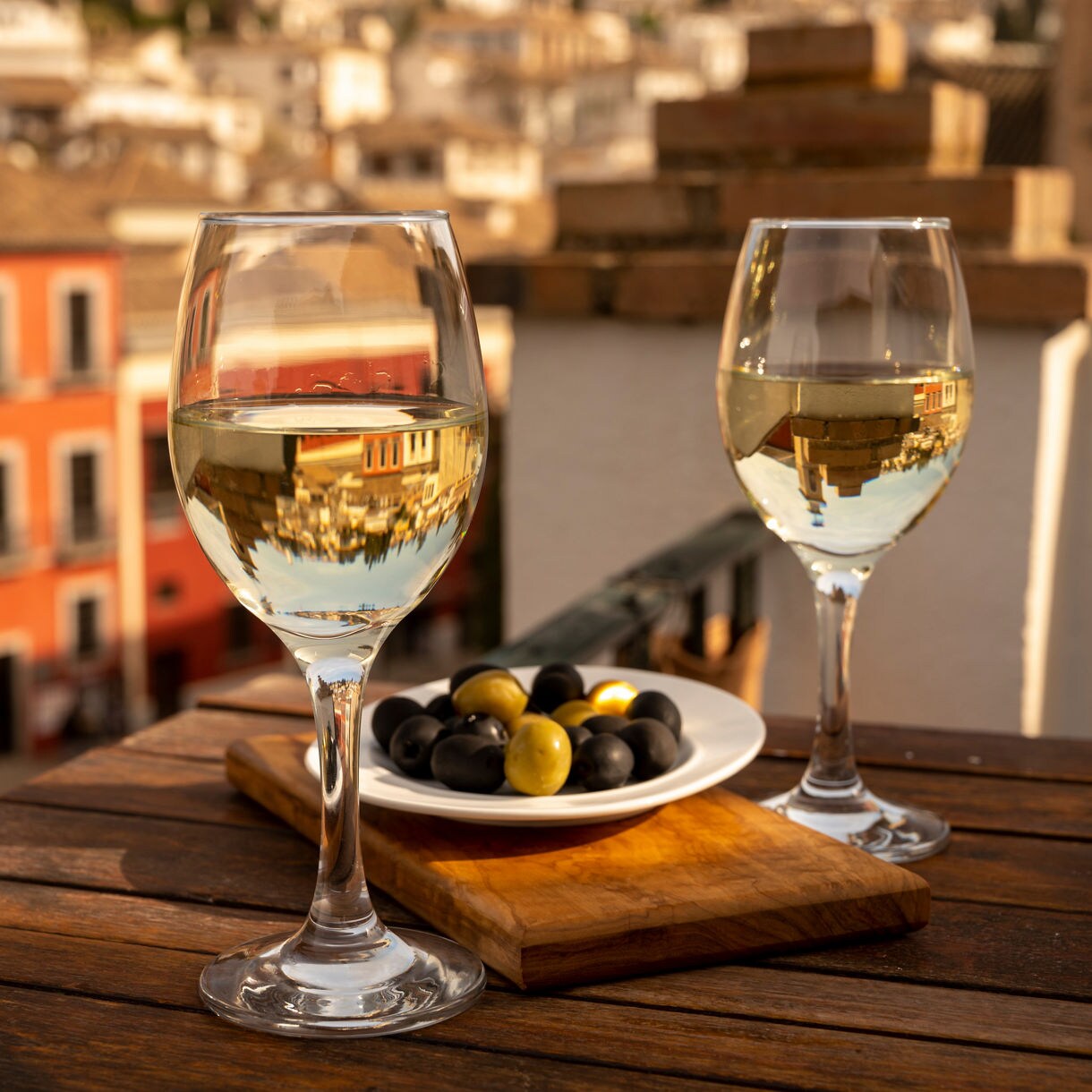 Two glasses of white wine and a plate of mixed olives on a wooden table overlooking a sun-drenched Spanish city square with colorful buildings below.