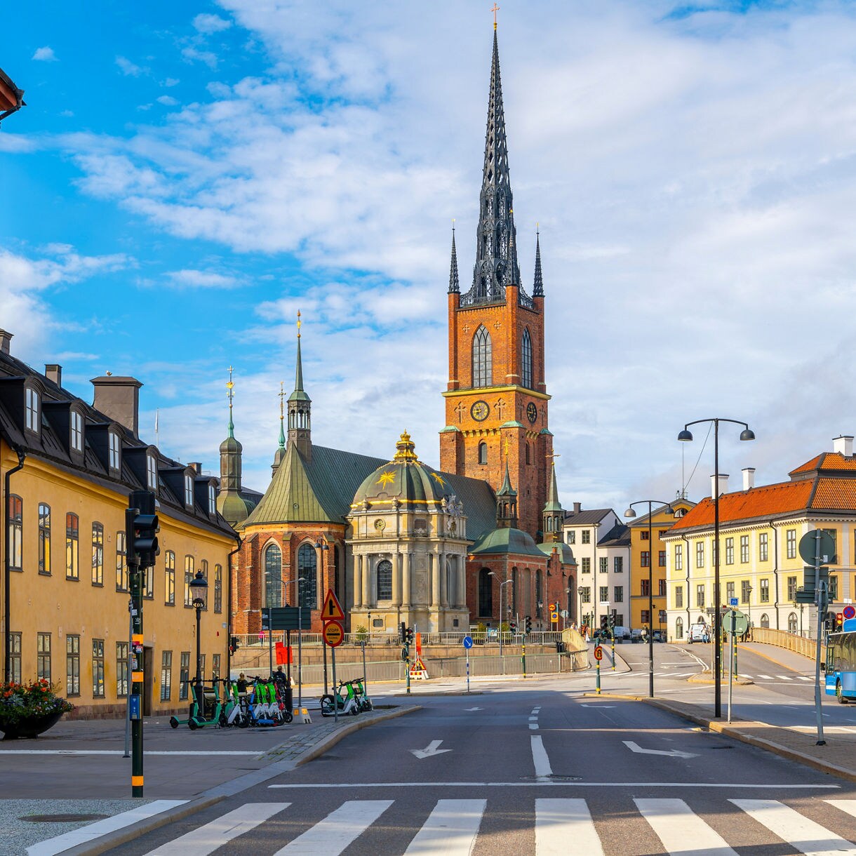 Street view leading to Riddarholmen Church in Stockholm, with its tall black spire rising above colorful historic buildings on a clear day.