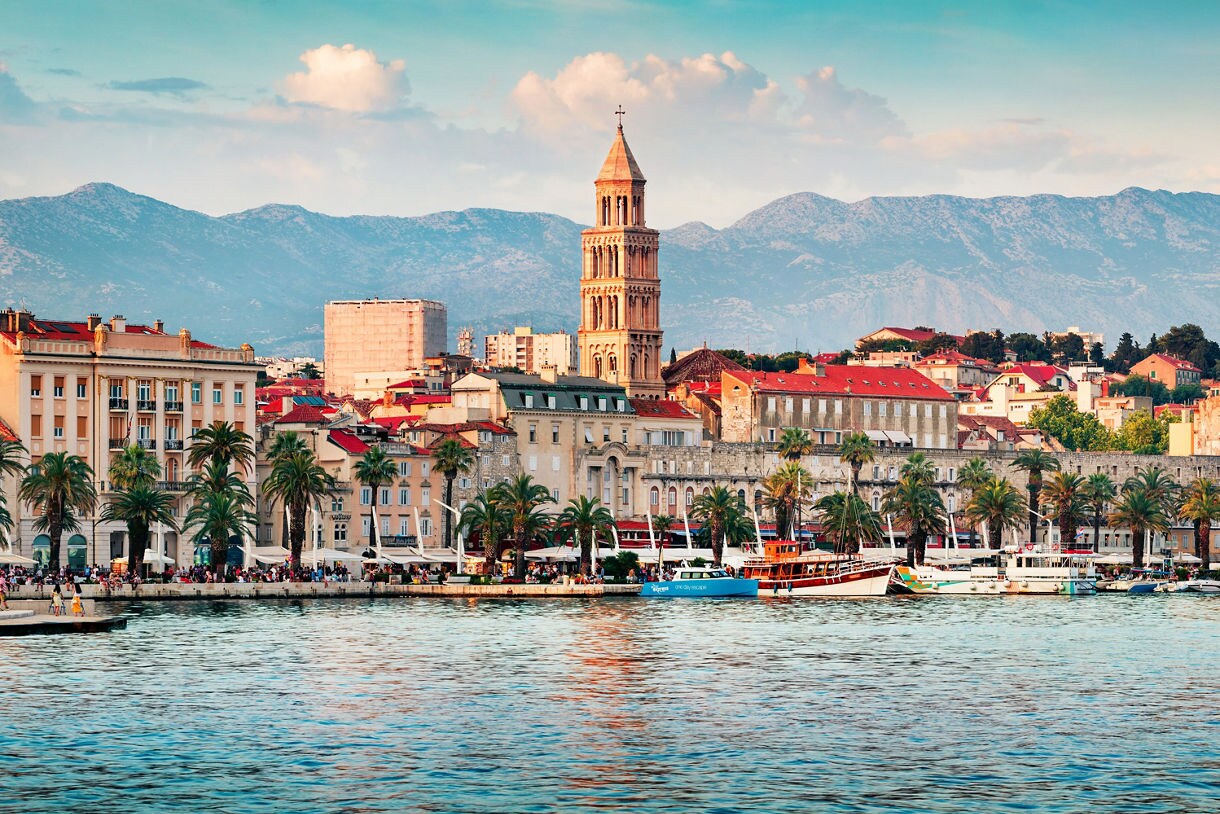 Waterfront view of Split, Croatia, showing a row of historic buildings with red-tiled roofs, palm trees along the promenade, boats in the harbor and mountains rising in the background.