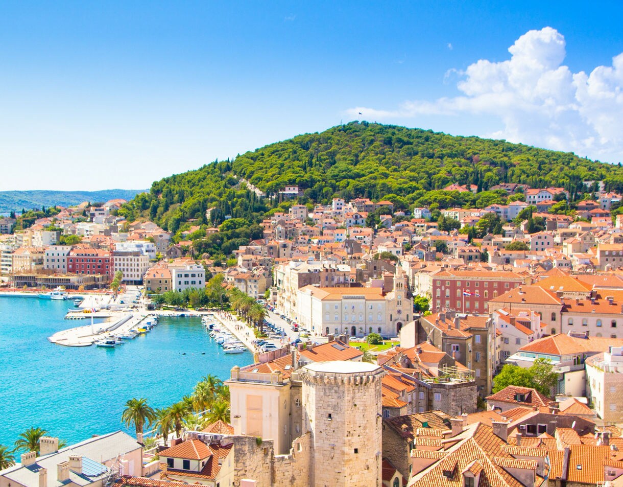A bright coastal view of Split, Croatia, showing terracotta rooftops, a waterfront lined with palm trees and clear blue water, with a forested hill rising behind the town.