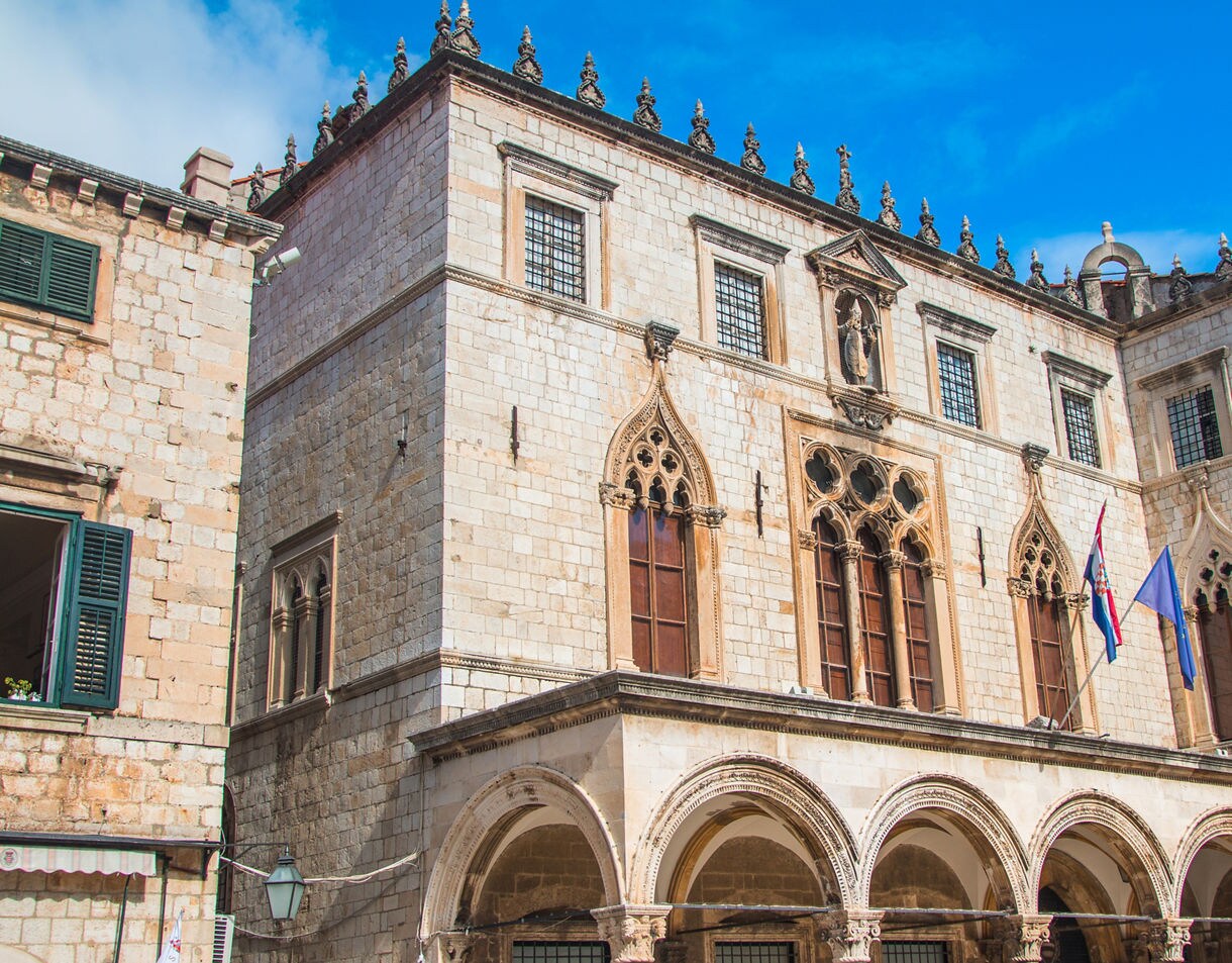 Exterior view of Sponza Palace in Dubrovnik, Croatia, featuring Gothic-Renaissance arches, ornate windows and stone detailing under a clear blue sky.