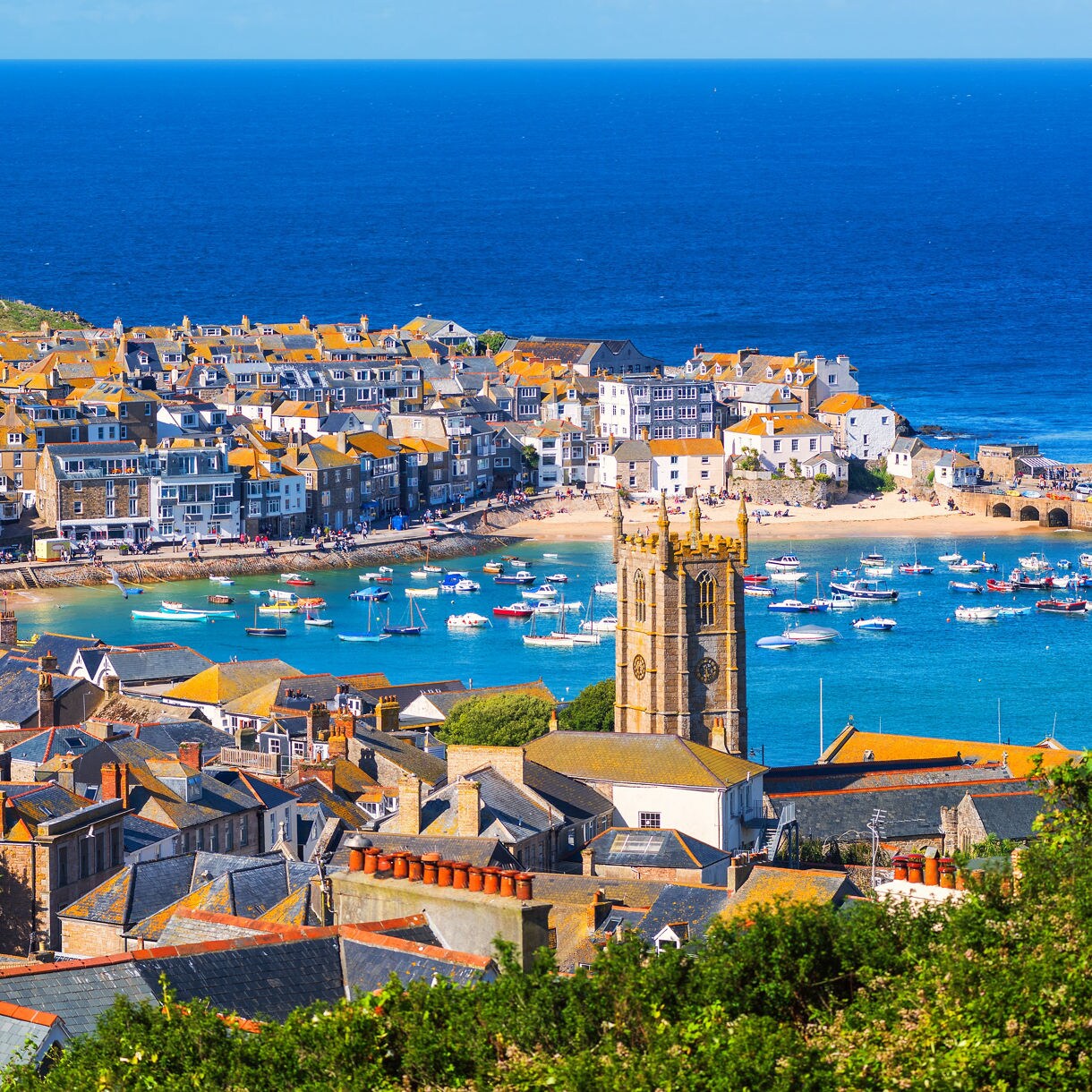 Aerial view of St Ives town in Cornwall with slate-roofed houses, a stone church tower, a busy harbor filled with boats and the deep blue ocean beyond.