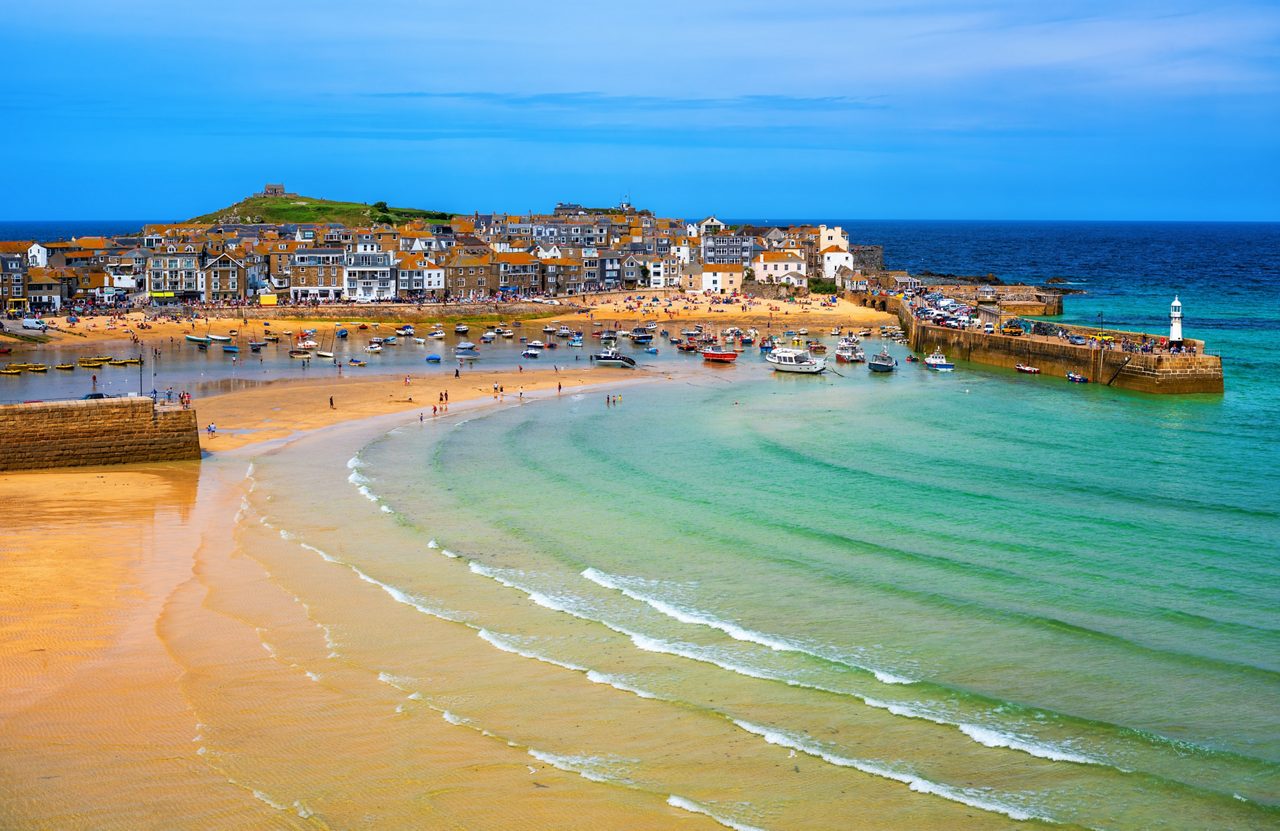 Colorful view of St Ives harbor with sandy beach, boats resting on the shore, clear turquoise water and crowds enjoying the summer sun.