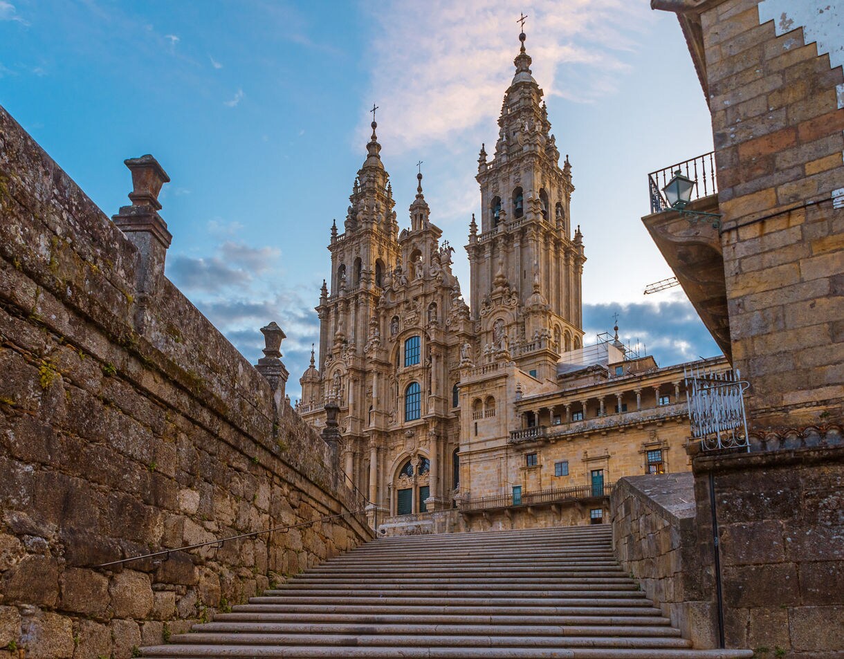 Wide stone staircase leading up to the ornate twin towers of Santiago de Compostela Cathedral, lit by soft early evening light with a blue and pink sky overhead.