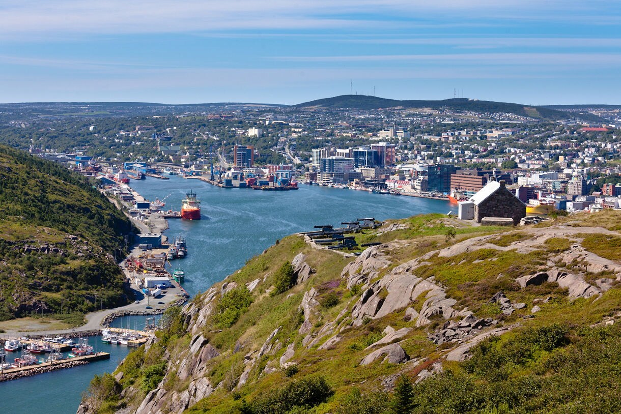 A wide view of St. John’s, Newfoundland, showing Signal Hill’s rocky slope in the foreground and the city’s harbor below with ships, docks and a dense spread of buildings under a clear blue sky.